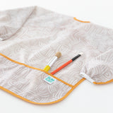 Bumkins Long-Sleeved Art Smock: Wander lays on a white background, with two paintbrushes in the front pocket.