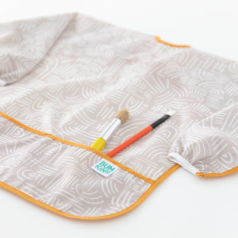 Bumkins Long-Sleeved Art Smock: Wander lays on a white background, with two paintbrushes in the front pocket.