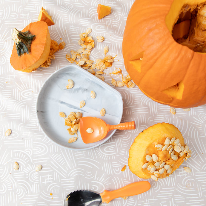 Pumpkin seeds, a partially carved pumpkin, carving tools, and a gray plate lay atop a beige mat with a white abstract pattern.