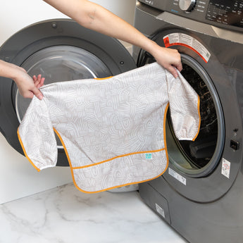 A person holds a Bumkins Long-Sleeved Art Smock: Wander in front of a front-loading washing machine.