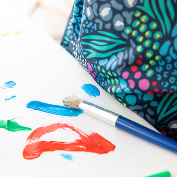Close-up of a child in a Bumkins Long-Sleeved Art Smock: Animal Prints next to a paintbrush and colorful strokes of paint on white paper.