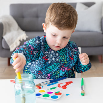 A young child wearing a Bumkins Long-Sleeved Art Smock: Animal Prints paints at a white table in front of a gray couch.