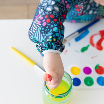 Close-up of a child wearing a Bumkins Long-Sleeved Art Smock: Animal Prints at a white table, dipping a brush in water next to a paint set.