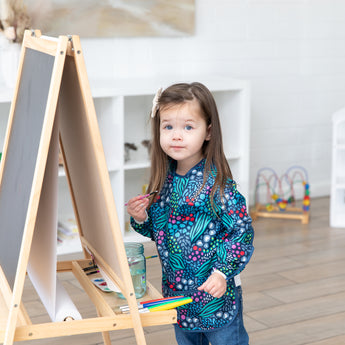 In a bright room, a young girl with long brown hair paints at an easel wearing a Bumkins Long-Sleeved Art Smock: Animal Prints.
