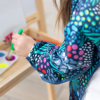 Close-up of a child painting with a green brush, wearing a Bumkins Long-Sleeved Art Smock: Animal Prints.