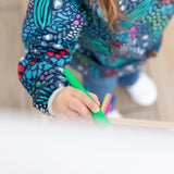 A child in a Bumkins Long-Sleeved Art Smock: Animal Prints draws with a green marker at an easel.