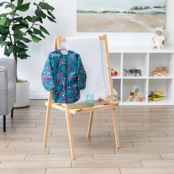 A Bumkins Long-Sleeved Art Smock: Animal Prints hangs from an easel in a bright room with toy-filled shelves.