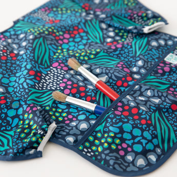 Bumkins Long-Sleeved Art Smock: Animal Prints rests on a white background with two paintbrushes in the pocket.