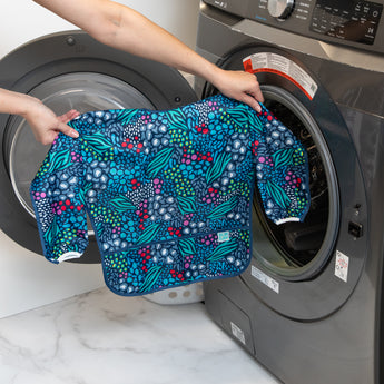 A person holds a Bumkins Long-Sleeved Art Smock: Animal Prints in front of an open front-loading washing machine.