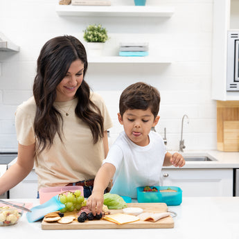 A woman and child in a kitchen making lunch, with the child reaching for fruit near a Bumkins Silicone Bento Box 3 Section: Blue Jelly.