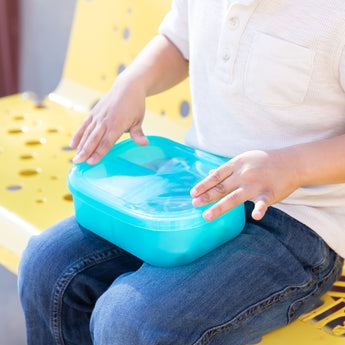 A child sits on a yellow bench, holding a Bumkins Silicone Bento Box 3 Section: Blue Jelly on their lap.
