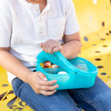 A child sits on a yellow bench, eagerly opening a Bumkins Silicone Bento Box 3 Section: Blue Jelly filled with fruits and snacks.