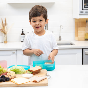 A smiling child makes a sandwich in a bright kitchen, surrounded by ingredients and a Bumkins Silicone Bento Box 3 Section: Blue Jelly.
