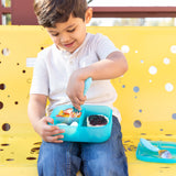 A child sits on a yellow bench, using a fork to eat from a Bumkins Silicone Bento Box 3 Section: Blue Jelly filled with snacks.
