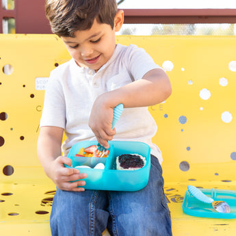A child sits on a yellow bench, using a fork to eat from a Bumkins Silicone Bento Box 3 Section: Blue Jelly filled with snacks.