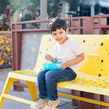 A young boy sitting on a yellow bench, smiling and holding a Bumkins Silicone Bento Box 3 Section: Blue Jelly filled with food.