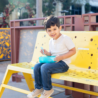A young boy sitting on a yellow bench, smiling and holding a Bumkins Silicone Bento Box 3 Section: Blue Jelly filled with food.