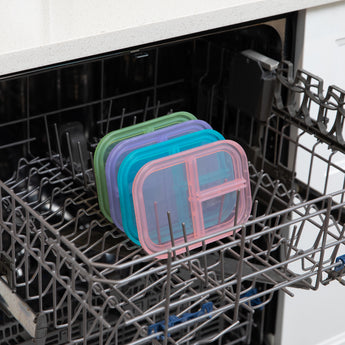 Four lids, including one from Bumkins Silicone Bento Box 3 Section: Green Jelly, sit in the top rack of an open dishwasher.