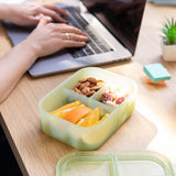 An open bento box from Bumkins Silicone Bento Box 3 Section 2 Pack: Aqua Jelly filled with snacks sits next to a person typing on a laptop.