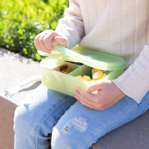 Child in a cream sweater opens a Bumkins Silicone Bento Box 3 Section (Aqua Jelly) outdoors on a sunny day.