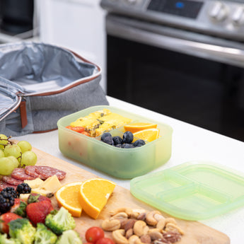 A bento box from Bumkins Lunchbox Legends 6-Piece Set: Motley Green Jelly sits on a kitchen counter by a lunch bag, fruit, veggies, & nuts.