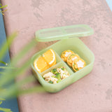 A bento box from Bumkins Lunchbox Legends 6-Piece Set: Motley Green Jelly with crackers, pasta salad, & orange slices sits on tan concrete.