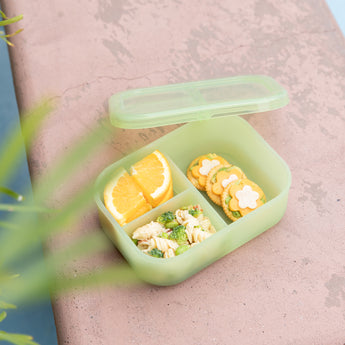 A bento box from Bumkins Lunchbox Legends 6-Piece Set: Motley Green Jelly with crackers, pasta salad, & orange slices sits on tan concrete.