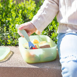 A child uses Bumkins Spoon + Fork: Sage to eat from a green bento box on a concrete bench by greenery.