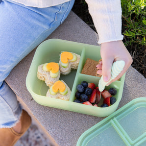 A child eats fruit and sandwiches from a Bumkins Silicone Bento Box 3 Section: Green Jelly with a fork outdoors.