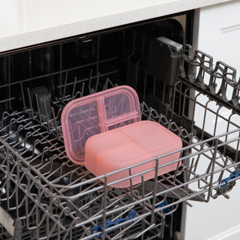 A bento box and lid from Bumkins Lunchbox Legends 6-Piece Set: Watercolor Pink Jelly sit in the top rack of an open dishwasher.
