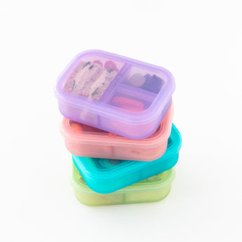A stack of colorful bento boxes filled with snacks, including a Bumkins Silicone Bento Box 3 Section: Purple Jelly, on a white background.