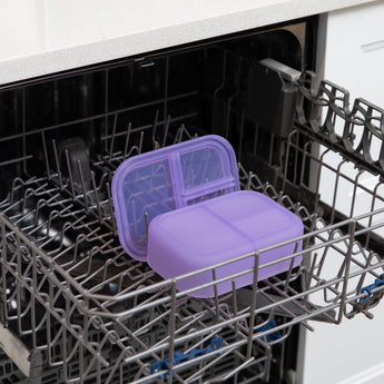 A bento box and lid from Bumkins Lunchbox Legends 6-Piece Set: Unicorns Purple Jelly sit in the top rack of an open dishwasher.