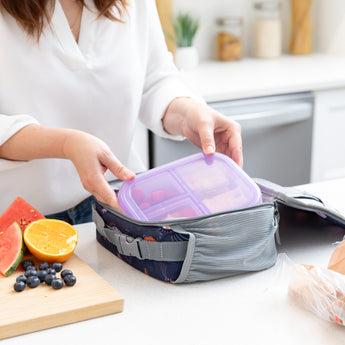A woman places a bento box from Bumkins Lunchbox Legends 6-Piece Set: Coastal Purple Jelly in a lunch bag on a kitchen counter.