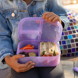 A child holds a Bumkins Silicone Bento Box 3 Section: Purple Jelly, filled with a sandwich, tomatoes, carrots, grapes, and blackberries.