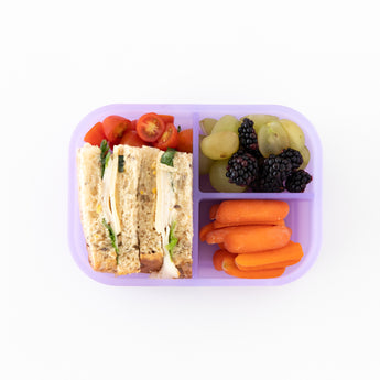 Bumkins Silicone Bento Box 3 Section: Purple Jelly holds a sandwich, grapes, blackberries, carrots, and cherry tomatoes on a white background.