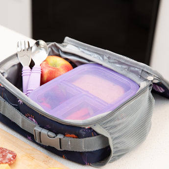 Bumkins Spoon + Fork: Lavender sit in an open lunch bag with an apple and a transparent purple bento box on a kitchen counter.