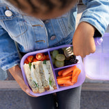 A child wearing a denim jacket with a Bumkins Silicone Bento Box 3 Section: Purple Jelly, filled with a sandwich, fruits, and veggies.