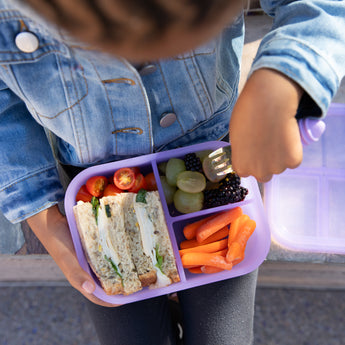 A child wearing a denim jacket with a Bumkins Silicone Bento Box 3 Section: Purple Jelly, filled with a sandwich, fruits, and veggies.