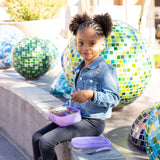 A curly-haired girl sits on a bench with a Bumkins Silicone Bento Box 3 Section: Purple Jelly, against colorful mosaic spheres.