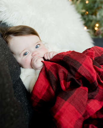 A blue-eyed baby lies on a couch with Little Unicorns red plaid Cotton Swaddle, a white pillow, and a Christmas tree behind.