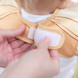Adult hands fasten the hook-and-loop closure of a Bumkins Ruffle Apron Bib: Belle around a child's neck.