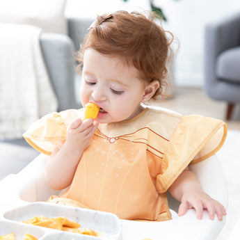 A toddler sits in a high chair, wearing a Bumkins Ruffle Apron Bib: Belle and eating from a gray plate in a bright room.