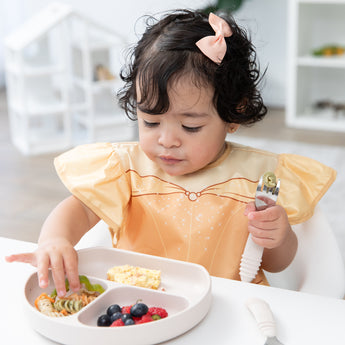 A toddler in a high chair wearing a Bumkins Ruffle Apron Bib: Belle holds a spoon and eats from a beige divided plate of pasta and berries.