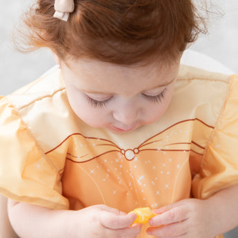 Close-up of a toddler with red hair in a Bumkins Ruffle Apron Bib: Belle looking down, holding an orange fruit.