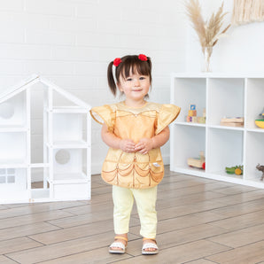 A young girl stands in a bright playroom, wearing a Bumkins Ruffle Apron Bib: Belle.