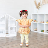 A young girl stands in a bright playroom, wearing a Bumkins Ruffle Apron Bib: Belle.