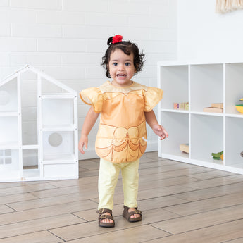 A girl in a ponytail smiles in a bright playroom, wearing a bib from Bumkins Snack In Character 4-Piece Set: Disney Princess Magic Belle.