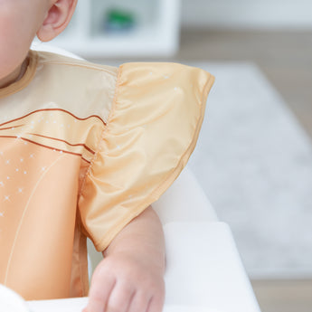 Close-up of a child in a high chair wearing a Bumkins Ruffle Apron Bib: Belle, with focus on the ruffle sleeve.