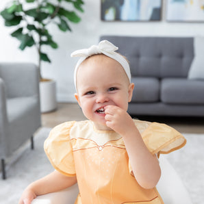 A girl in a bow smiles, wearing a bib from Bumkins Snack In Character 4-Piece Set: Disney Princess Magic Belle.