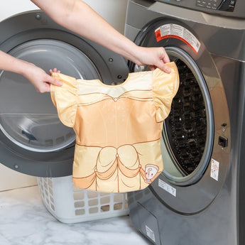 A person holds a Bumkins Ruffle Apron Bib: Belle in front of an open front-loading washing machine.